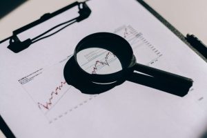 Symbolbild: Eine Lupe auf einem Ausdruck mit Börsenkursen symbolisiert die Prüfung von Finanzanlagenvermittlern