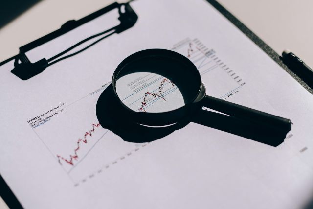 Symbolbild: Eine Lupe auf einem Ausdruck mit Börsenkursen symbolisiert die Prüfung von Finanzanlagenvermittlern