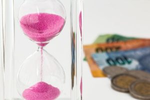 Symbolbild: Eine Sanduhr neben einer Hand mit Geldscheinen symbolisiert die einzuhaltenden Fristen, die im Gewinnabführungsvertrag verankert sind.